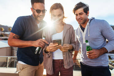 Three young friends at rooftop party looking at the smart phone together. Group of young men and woman standing and using mobile phone. - JLPSF03186