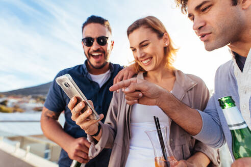 Eine Gruppe von Freunden auf einer Dachterrassenparty schaut auf ihr Smartphone und lächelt. Junge Männer und eine Frau stehen auf einer Dachterrasse und benutzen ein Mobiltelefon. - JLPSF03187