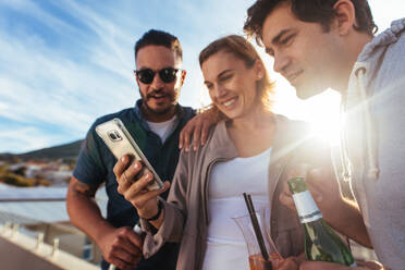 Gruppe von Männern und Frauen auf einer Party, die sich ein Video auf dem Mobiltelefon ansehen. Drei junge Freunde stehen zusammen und schauen auf ihr Smartphone. - JLPSF03188