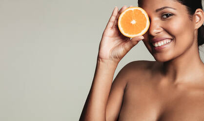 Nahaufnahme einer schönen jungen Frau ohne Hemd, die ein Stück Orange vor ihrem Auge hält, während sie vor einem grauen Hintergrund steht. - JLPSF03228