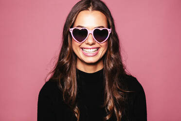 Porträt der schönen jungen Frau mit herzförmiger Sonnenbrille und lächelnd gegen rosa Hintergrund. kaukasischen Mode-Modell posiert mit Brille. - JLPSF03276