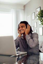 Unbekümmerter junger Mann hört Musik, während er allein zu Hause sitzt. Gut aussehender junger schwarzer Mann, der fröhlich lächelt, während er mit Kopfhörern eine Wiedergabeliste auf seinem Laptop anhört. - JLPSF03300