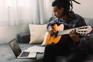 Black male musician learning guitar lessons online. Young guitarist holding a guitar while watching a music tutor on his laptop. Man studying music at home during quarantine. - JLPSF03303