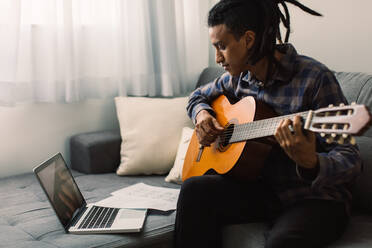 Schwarzer männlicher Musiker, der online Gitarrenunterricht nimmt. Junger Gitarrist, der eine Gitarre in der Hand hält, während er einen Musiklehrer auf seinem Laptop beobachtet. Mann, der während der Quarantäne zu Hause Musik studiert. - JLPSF03303