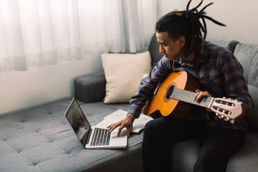 Junger Musiker, der online Gitarrenunterricht nimmt. Junger Gitarrist, der eine Gitarre in der Hand hält, während er einen Musiklehrer auf seinem Laptop beobachtet. Mann, der während der Quarantäne zu Hause Musik studiert. - JLPSF03304