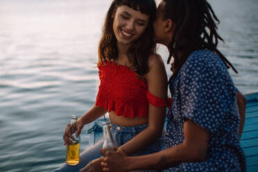 Interracial couple enjoying a romantic moment together duiring a vacation. Two affectionate young lovers being playful with each other while sitting together on a dock and holding beer bottles. - JLPSF03319