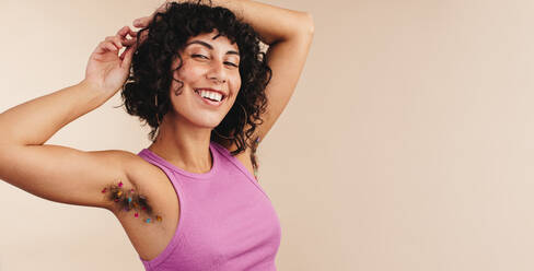 Cheerful woman smiling at the camera while showing off her confetti-decorated underarm hair. Happy young woman making the choice not to shave. Body confident young woman embracing her natural body. - JLPSF03370