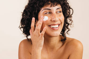 Happy young woman smiling cheerfully while applying moisturising cream on her face. Attractive young woman treating her facial skin with a nourishing beauty product. - JLPSF03391