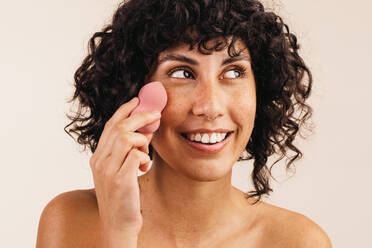 Pretty young woman looking away with a smile while using a makeup sponge on her face. Happy young woman using a beauty blender to apply makeup foundation on her face. - JLPSF03405
