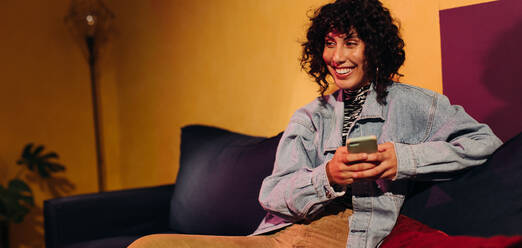 Glückliche junge Frau, die fröhlich lächelt, während sie zu Hause ein Smartphone benutzt. Unbekümmerte junge Frau, die eine Textnachricht sendet, während sie sich auf einer Couch in einem Haus entspannt. - JLPSF03412