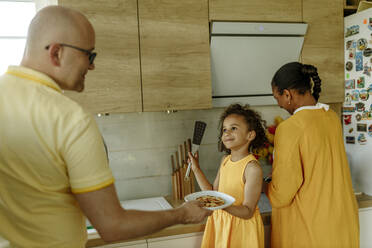 Lächelndes Mädchen gibt dem Vater einen Teller, der neben der Mutter in der Küche steht - VIVF00016
