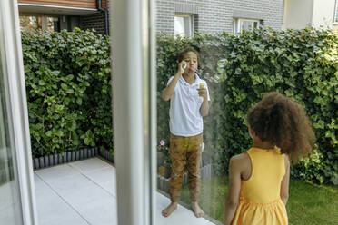 Girl looking at brother blowing soap bubbles on terrace - VIVF00054