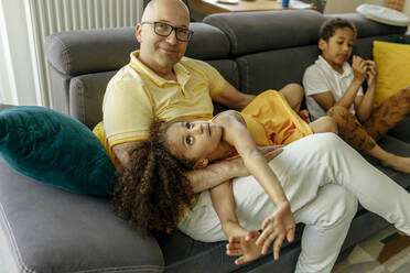 Girl lying on father's lap by brother on sofa at home - VIVF00062