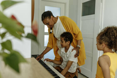 Mädchen betrachtet Mutter, die ihrem Sohn zu Hause das Klavierspielen beibringt - VIVF00068