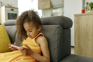 Mädchen mit lockigem Haar, das auf dem Sofa zu Hause ein Video über das Mobiltelefon ansieht - VIVF00076