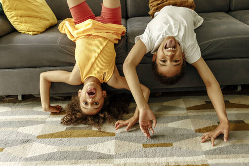 Mädchen und Junge strecken Zunge heraus und genießen kopfüber auf dem Sofa zu Hause - VIVF00078