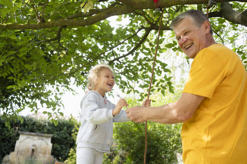 Glücklicher älterer Mann mit Enkelin, die mit einem Seil im Garten schaukeln - SVKF00584