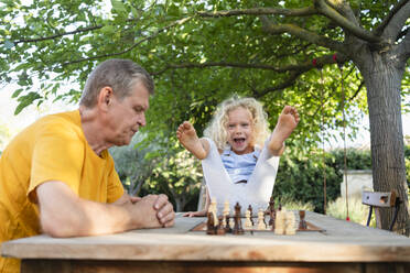 Glückliches Mädchen, das sich am Großvater erfreut, der auf einem Tisch im Garten Schach spielt - SVKF00603