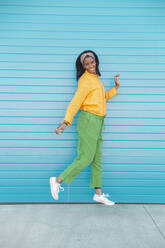 Smiling young woman jumping in front of blue wall - PCLF00066