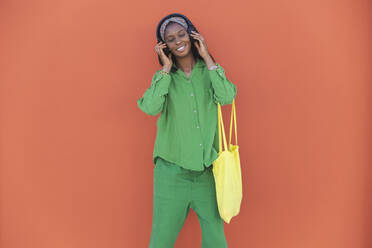 Junge Frau genießt Musik über Kopfhörer vor einer orangefarbenen Wand - PCLF00078