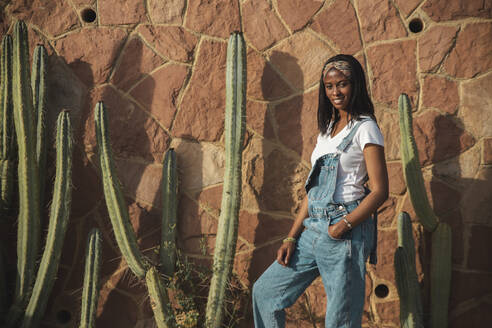 Lächelnde junge Frau steht vor einer Mauer mit Kaktus an einem sonnigen Tag - PCLF00090