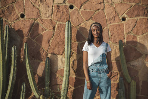 Selbstbewusste junge Frau vor einer Wand mit Kaktuspflanze - PCLF00091