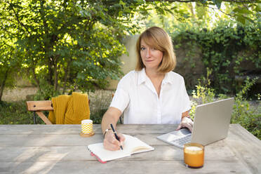Freiberufler macht sich Notizen, sitzt mit Laptop am Tisch im Garten - SVKF00610
