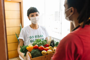 Box of vegan food being delivered to a home. Young delivery woman with a face mask delivering a box of fresh vegetables to a young man at his home. Home delivery during the covid-19 pandemic. - JLPSF03429