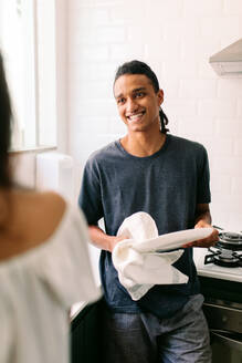 Mann beim Abwaschen mit seiner Freundin. Ausschnitt eines gutaussehenden jungen Mannes, der seine Freundin anlächelt, während er in der Küche das Geschirr abwischt. Ein Paar bei der Hausarbeit zu Hause. - JLPSF03439