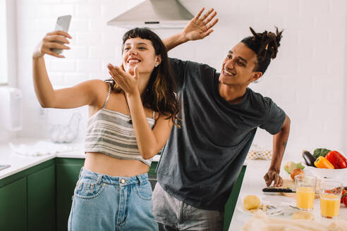 Carefree young couple smiling happily while video calling their friends in the kitchen. Happy young couple greeting their friends on a video call. Couple cooking together at home. - JLPSF03460