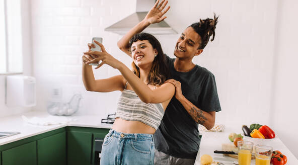 Cheerful young couple smiling happily while taking a selfie in their kitchen. Happy young couple capturing their cooking moments at home. Young couple cooking together. - JLPSF03462