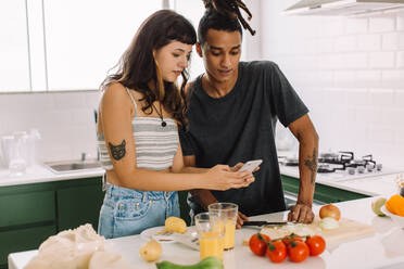 Junges Paar, das ein Online-Rezept auf einem Smartphone verfolgt. Romantisches junges Paar, das im Internet nach Rezepten sucht, während es in der Küche sein Essen zubereitet. Paar, das zusammen kocht. - JLPSF03464