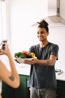 Young couple taking a photo of their food at home. Happy young man smiling cheerfully while holding a plate of raw fresh food in the kitchen. Food bloggers recording content for their cooking blog. - JLPSF03468