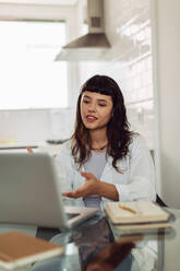 Lächelnde junge Geschäftsfrau, die an einem virtuellen Meeting teilnimmt, während sie allein an ihrem Schreibtisch im Home Office sitzt. Junge Geschäftsfrau, die einen Videoanruf auf ihrem Laptop entgegennimmt. Frau, die von zu Hause aus arbeitet. - JLPSF03470