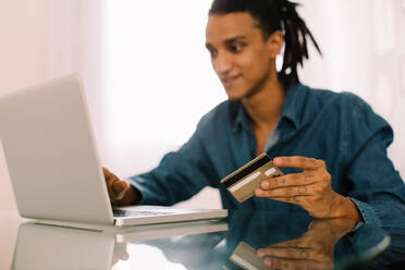 Man shopping online at home during the day. Handsome young man using a credit card and a laptop to make online purchases while sitting alone indoors. - JLPSF03476