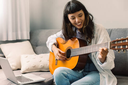 Glückliche Musikerin, die an ihrem Laptop Gitarrenunterricht gibt. Junge Gitarristin, die per Videoanruf online Musikunterricht gibt. Frau, die während einer Quarantäne zu Hause Musikunterricht gibt. - JLPSF03478