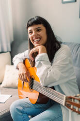 Female musician holding a guitar at home. Happy young female guitarist smiling cheerfully while sitting alone at home during the day. - JLPSF03480