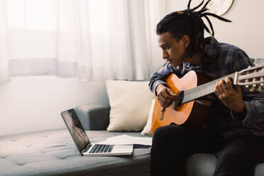 Afroamerikanischer Musiker, der online Gitarrenunterricht nimmt. Junger Gitarrist, der eine Gitarre in der Hand hält und einen Musiklehrer auf seinem Laptop beobachtet. Mann, der während der Quarantäne zu Hause Musik studiert. - JLPSF03481