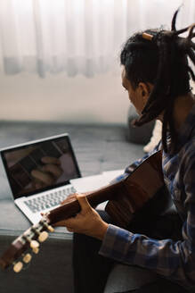 Junger Gitarrist, der an seinem Laptop Online-Gitarrenunterricht nimmt. Junger männlicher Musiker, der eine Gitarre hält, während er per Videoanruf eine Gitarrenstunde verfolgt. Mann, der während einer Quarantäne zu Hause Musik studiert. - JLPSF03482