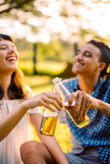 Lächelndes junges Paar, das im Freien mit Bierflaschen anstößt. Zwei romantische junge Verliebte, die tagsüber in einem Park sitzen und einen Moment zusammen feiern. - JLPSF03492