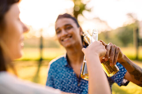 Lächelndes junges Paar, das einen Moment im Freien feiert. Zwei romantische junge Verliebte stoßen fröhlich mit Bierflaschen an, während sie tagsüber zusammen in einem Park sitzen. - JLPSF03496