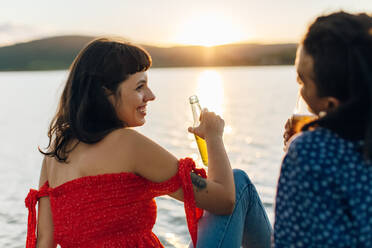 Lächelndes junges gemischtrassiges Paar, das sich im Urlaub bei einem Bier entspannt. Zwei romantische junge Liebende, die sich gemütlich unterhalten, während sie mit Bierflaschen in der Hand am Meer sitzen. - JLPSF03503