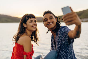 Glückliches junges gemischtrassiges Paar, das im Urlaub einen Moment festhält. Zwei romantische junge Liebende, die fröhlich lächeln, während sie tagsüber gemeinsam Selfies am Meer machen. - JLPSF03506