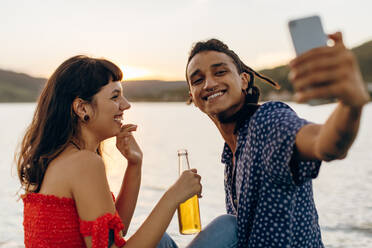 Cute young interracial couple capturing a moment while on a vacation. Two romantic young lovers smiling cheerfully while taking a selfie together at the sea during the day. - JLPSF03507