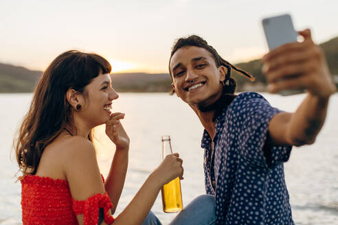 Nettes junges interrassisches Paar, das im Urlaub einen Moment festhält. Zwei romantische junge Liebende lächeln fröhlich, während sie tagsüber ein Selfie am Meer machen. - JLPSF03507