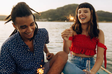 Fröhliches junges Paar, das im Urlaub mit bengalischen Lichtern spielt. Zwei romantische junge Liebende lachen fröhlich, während sie bengalische Lichter halten und zusammen am Meer sitzen. - JLPSF03511