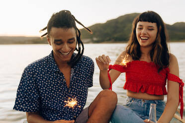 Glückliches junges Paar, das sich im Urlaub mit bengalischen Lichtern vergnügt. Zwei romantische junge Liebende lächeln fröhlich, während sie bengalische Lichter halten und zusammen am Meer sitzen. - JLPSF03512