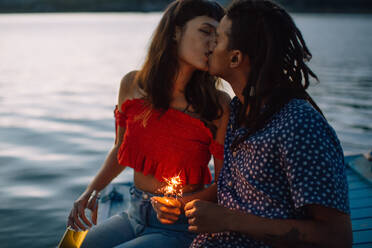 Romantisches junges Paar, das im Urlaub einen leidenschaftlichen Moment miteinander teilt. Zwei zärtliche junge Liebende, die sich küssen, während sie zusammensitzen und bengalische Lichter am Meer halten. - JLPSF03514