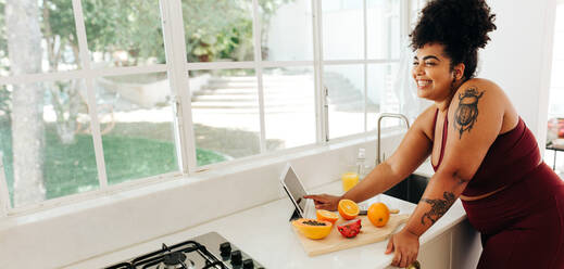 Weitwinkelaufnahme einer attraktiven Frau, die in der Küche steht und ein Rezept auf einem digitalen Tablet liest. Eine Frau mit Übergröße in Sportkleidung benutzt einen Tablet-PC, während sie zu Hause einen gesunden Fruchtsaft herstellt. - JLPSF03527