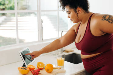 Healthy woman standing and reading recipe on digital tablet at kitchen. Body positive female in sports clothing using tablet pc in kitchen while preparing fresh fruit juice. - JLPSF03528